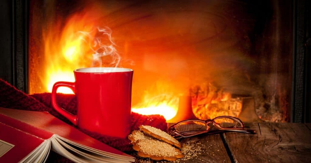 A hot chocolate cup next to a fireplace, accompanied by a book and a biscuit.